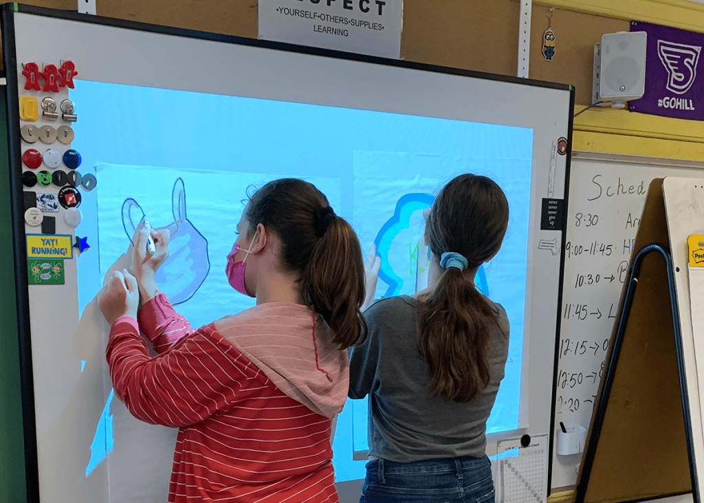 Two students trace Marne debris items on a projector display in their classroom.