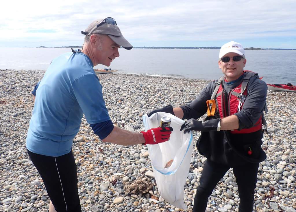Two men pick up trash on the beach and put it in a trash bag.