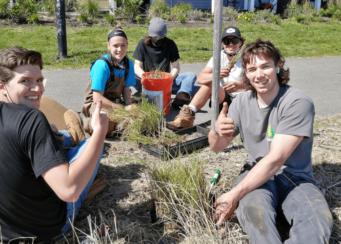 Teens volunteer for salt marsh restoration, sitting on the ground and smiling.